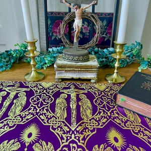 May include: A purple and gold fabric altar cloth with a cross and angels. A small wooden crucifix with a figure of Jesus Christ is on a gold stand in front of the cloth. Two gold candle holders with white candles are on either side of the crucifix.