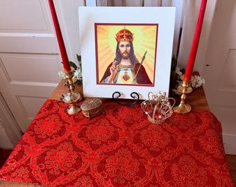 May include: A religious display featuring a framed image of Jesus Christ, flanked by two red candles in gold candle holders. A red patterned runner and a decorative crown are also present.