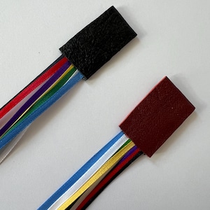 May include: Two leather bookmarks with colorful ribbon tassels. The bookmark on the top is black leather and the bookmark on the bottom is burgundy leather.