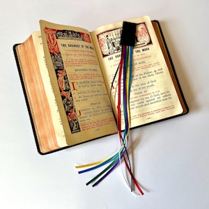 K&ouml;nnte beinhalten: Ein offenes Buch mit einem schwarzen Lesezeichen und mehreren bunten B&auml;ndern. Das Buch ist auf einer Seite mit dem Text "The Ordinary of the Mass" und "The Mass" ge&ouml;ffnet.