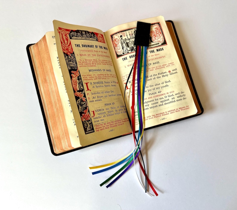 K&ouml;nnte beinhalten: Ein offenes Buch mit einem schwarzen Lesezeichen und mehreren bunten B&auml;ndern. Das Buch ist auf einer Seite mit dem Text "The Ordinary of the Mass" und "The Mass" ge&ouml;ffnet.