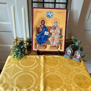 May include: A gold table runner with a repeating cross pattern. Two white candles are in gold candle holders. A framed icon of the Holy Trinity with the text "HOLY TRINITY" is on the table. Two small framed icons of saints are on the table.