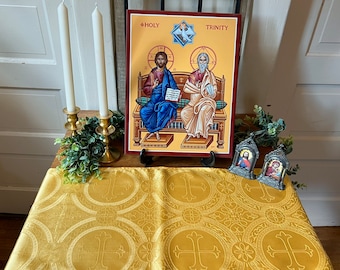 May include: A gold table runner with a repeating cross pattern. Two white candles are in gold candle holders. A framed icon of the Holy Trinity with the text "HOLY TRINITY" is on the table. Two small framed icons of saints are on the table.