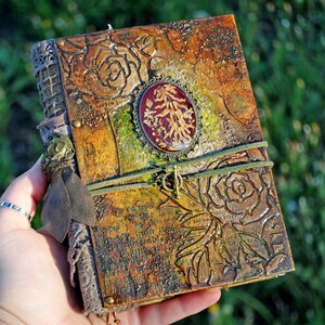 Puede incluir: Un diario artesanal con una apariencia texturizada y envejecida. La cubierta presenta rosas en relieve, un óvalo central con follaje seco y un cierre de cordón verde. La paleta de colores incluye marrones, verdes y acentos dorados, creando una estética vintage.