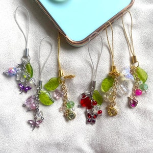 May include: Five colorful phone charms with green leaves, butterflies, and beads. The charms are attached to strings and are hanging from a white surface.
