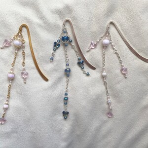 May include: Three silver and gold bookmark pendants with dangling chains of beads and charms. The charms include pink and blue butterflies, clear and opaque beads, and a larger, round, opaque bead.