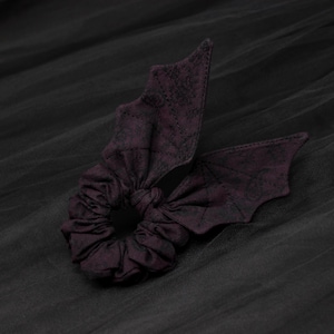 May include: A dark purple fabric hair scrunchie featuring two attached bat wing shapes. The fabric has a subtle, mottled texture throughout the circular elastic band and the pointed, stitched wing extensions that extend from the center.