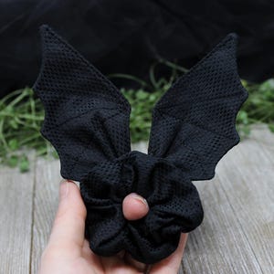 May include: A black hair scrunchie featuring two attached fabric bat wings. The accessory is made from a textured, mesh-like fabric with a consistent matte black finish throughout the entire piece, including the wing structures and the elastic band.