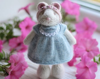May include: A small knitted mouse wearing a light blue dress with a white lace trim and a pink headband with flowers. The mouse is standing on a white wooden surface with pink flowers.