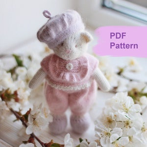 May include: A knitted white cat doll wearing a pink outfit and a purple beret. The doll is surrounded by white cherry blossoms on a white wooden surface. The text "PDF Pattern" is written in pink on a white background.
