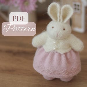 Könnte beinhalten: Ein gestricktes Spielzeughäschen mit langen Ohren, das eine cremefarbene Strickjacke mit kleinen Knöpfen über einem blassrosa Rock mit dezentem Rautenmuster trägt. Das Bild enthält den Text PDF Pattern in einem weißen Kasten links.