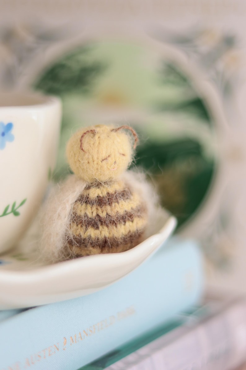 Pode incluir: Uma figura de abelha em miniatura feita &agrave; m&atilde;o, com uma cabe&ccedil;a amarela e um corpo listrado marrom e amarelo, repousando sobre um pires branco. O pires est&aacute; em cima de um livro azul claro, com uma x&iacute;cara de ch&aacute; com detalhes florais azuis ao fundo.
