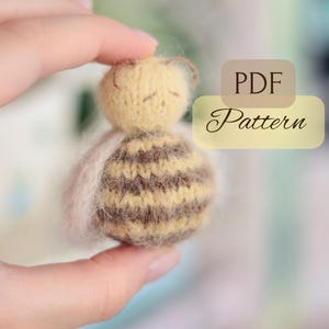 Pode incluir: Uma pequena boneca em forma de abelha, feita &agrave; m&atilde;o, com cabe&ccedil;a amarela e corpo listrado marrom e bege. A imagem inclui o texto "PDF Pattern". A boneca &eacute; segurada em uma m&atilde;o.