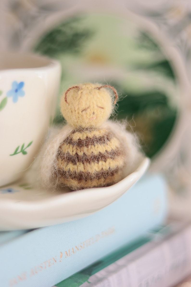 Pode incluir: Uma pequena figura de abelha de malha feita &agrave; m&atilde;o com listras marrons e amarelas, repousando sobre um pires branco. Uma x&iacute;cara de ch&aacute; floral ao fundo, com um livro azul claro vis&iacute;vel abaixo. Um item decorativo.