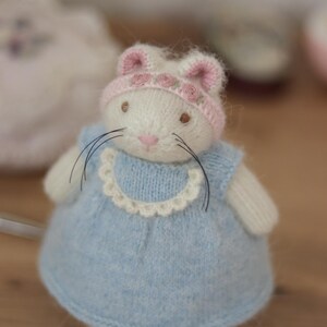 May include: A handmade white knitted bunny doll wearing a light blue dress with a white lace collar and a pink hat with rose accents. The doll has black whiskers and brown eyes. A charming, whimsical toy.