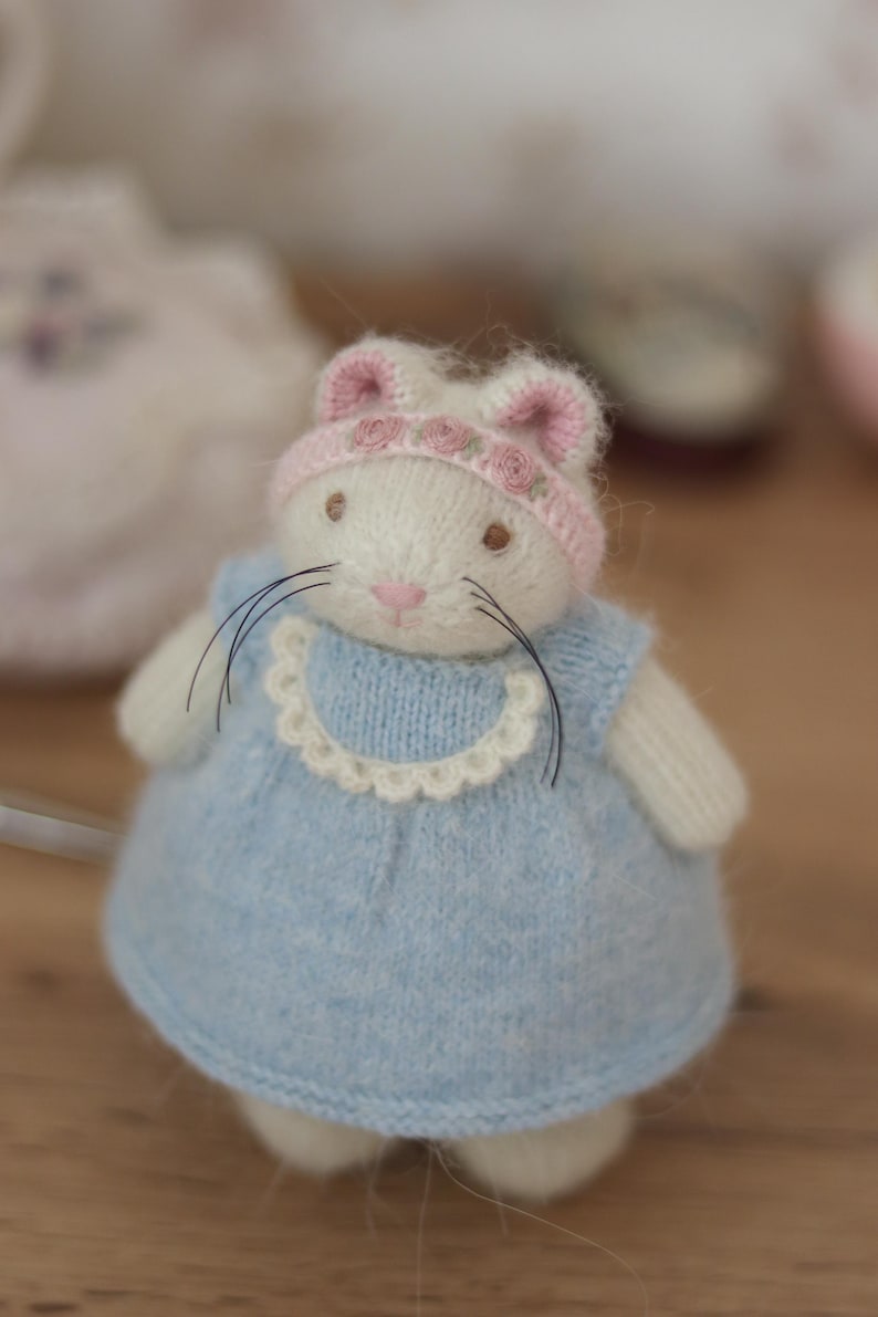 May include: A handmade white knitted bunny doll wearing a light blue dress with a white lace collar and a pink hat with rose accents. The doll has black whiskers and brown eyes. A charming, whimsical toy.