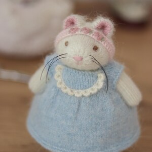 May include: A handmade white bunny doll wearing a light blue dress with a white scalloped collar and a pink hat adorned with small pink roses. The bunny has long black whiskers and a pink nose. The doll is made of soft, knitted material.