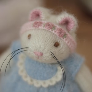 May include: A handmade white bunny doll with a pink nose and brown eyes. The bunny wears a light blue dress with a scalloped collar and a pink and white hat decorated with small pink roses. The doll has long black whiskers.
