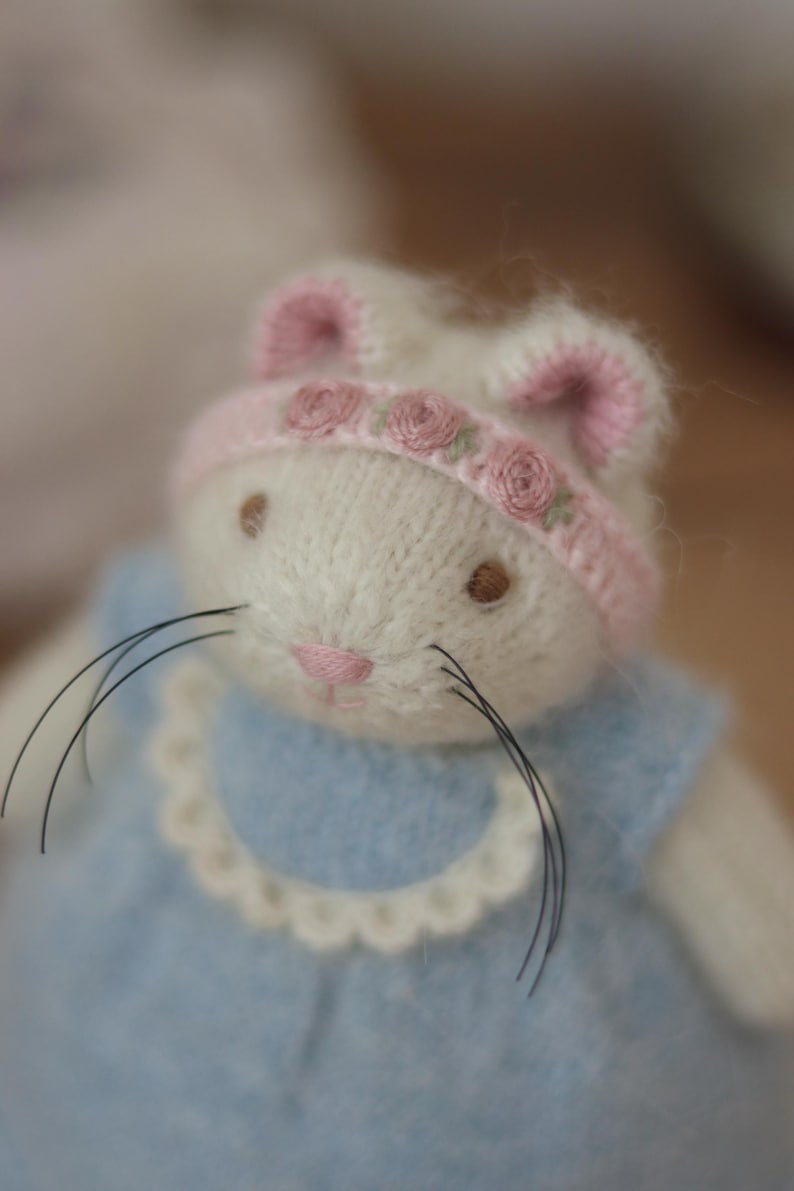 May include: A handmade white bunny doll with a pink nose and brown eyes. The bunny wears a light blue dress with a scalloped collar and a pink and white hat decorated with small pink roses. The doll has long black whiskers.