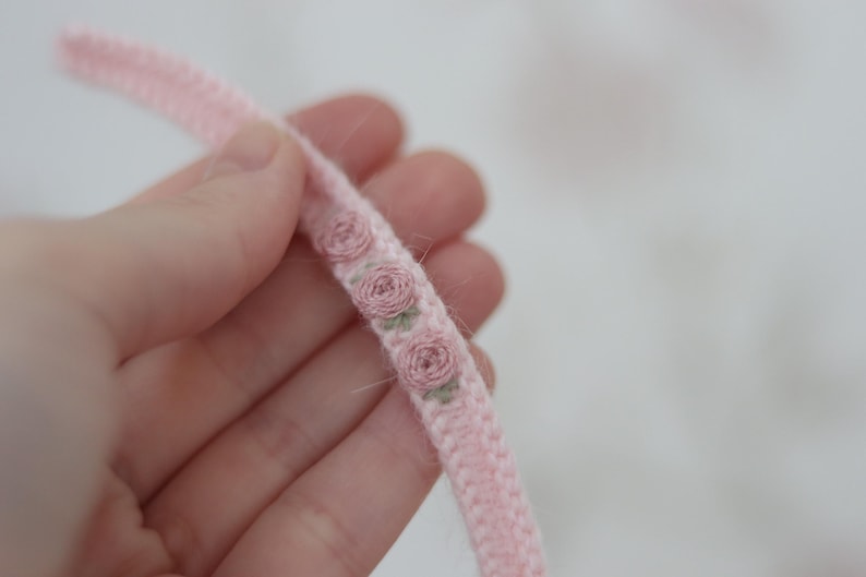 May include: A close-up of a pink, braided trim with embroidered rose details. The trim is held in a hand, showcasing the delicate floral pattern and soft texture. The roses are in shades of pink and green.