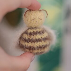 Pode incluir: Pequena boneca de abelha feita &agrave; m&atilde;o, com cabe&ccedil;a amarela e corpo listrado marrom e amarelo. A boneca tem olhos fechados e pequenas antenas marrons. A boneca &eacute; segurada em uma m&atilde;o.