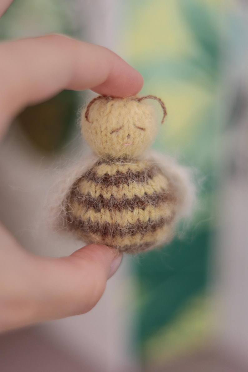 Pode incluir: Pequena boneca de abelha feita &agrave; m&atilde;o, com cabe&ccedil;a amarela e corpo listrado marrom e amarelo. A boneca tem olhos fechados e pequenas antenas marrons. A boneca &eacute; segurada em uma m&atilde;o.