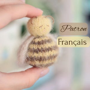 Può includere: Una piccola bambola fatta a mano a forma di ape, con testa gialla e corpo a righe marrone e beige. La bambola ha un alone sfocato di colore chiaro. Il testo "Patron Français" è sullo sfondo.