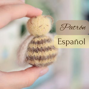 以下が含まれることがあります： 黄色い頭と茶色とベージュのストライプの体を持つ、手作りのミニチュアの蜂の人形。人形は目を閉じており、手に持たれています。画像には「Patrón Español」というテキストが含まれています。