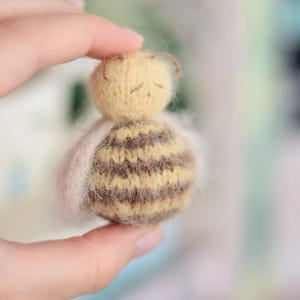 Pode incluir: Uma boneca de abelha em miniatura feita &agrave; m&atilde;o. A boneca tem uma cabe&ccedil;a amarela com pequenas antenas marrons e um corpo listrado marrom e amarelo. A boneca &eacute; segurada em uma m&atilde;o, com um material macio, branco e fofo ao redor do corpo.