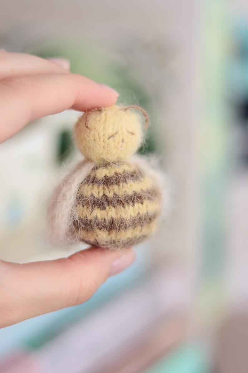 Pode incluir: Uma boneca de abelha em miniatura feita &agrave; m&atilde;o. A boneca tem uma cabe&ccedil;a amarela com pequenas antenas marrons e um corpo listrado marrom e amarelo. A boneca &eacute; segurada em uma m&atilde;o, com um material macio, branco e fofo ao redor do corpo.