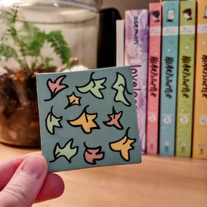 May include: A small, square canvas painted with a pattern of colorful leaves in shades of yellow, orange, and green on a teal background.