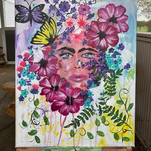 Pode incluir: Pintura abstrata em tela com o rosto de uma mulher rodeado por flores e borboletas coloridas. A obra utiliza uma paleta de rosas, roxos, azuis e verdes, com pinceladas vis&iacute;veis e um aspeto texturizado. A pintura &eacute; exibida num cavalete de madeira.