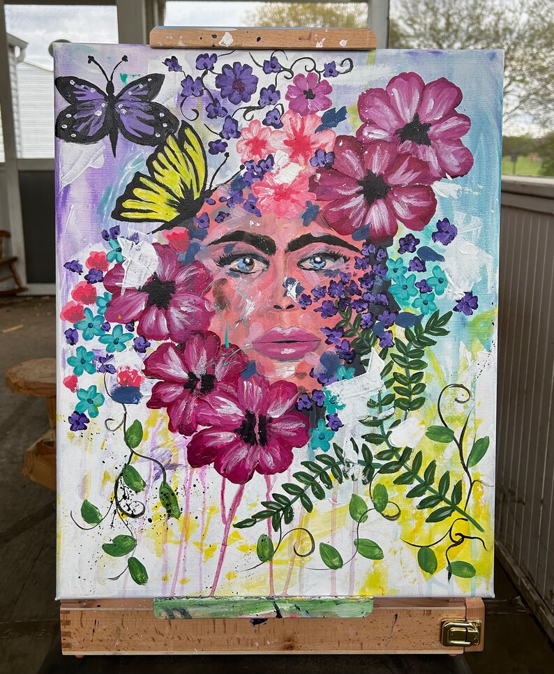 Pode incluir: Pintura abstrata em tela com o rosto de uma mulher rodeado por flores e borboletas coloridas. A obra utiliza uma paleta de rosas, roxos, azuis e verdes, com pinceladas vis&iacute;veis e um aspeto texturizado. A pintura &eacute; exibida num cavalete de madeira.