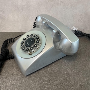 May include: Silver rotary dial telephone with a black cord. The phone features a circular dial with numbers and a central recall button. The handset is silver and rests on the base. The phone is a vintage style.