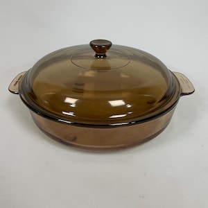 May include: A round, amber-coloured glass casserole dish with a matching transparent lid. The dish features integrated side handles and a central knob on the lid. The glass is translucent, allowing for a clear view of the interior base and sides.