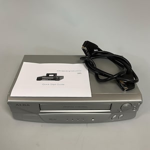 May include: Silver Alba VHS Hi-Fi VCR with a black cable and a quick start guide. The VCR has a remote control feature and several buttons and ports. The guide has a diagram of the VCR.