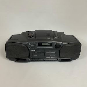 May include: A dark grey Philips boombox. The stereo has two speakers, a CD player, and a cassette deck. The boombox features a digital display and various control knobs and buttons. The text "TURBO BASS" is visible on the front.