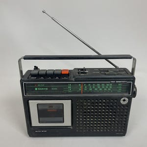 May include: A black Sanyo M 2408L portable radio cassette player with a silver cassette door, a black carrying handle, and a long, thin, extendable metal aerial. The front panel features a green radio frequency display, speaker grille, and buttons.
