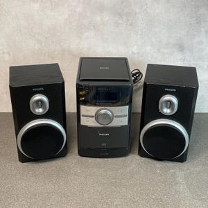 May include: A black Philips stereo system with two speakers. The stereo has a CD player and a digital display. The speakers have silver accents and a circular design. The system is on a neutral background.