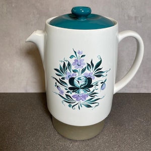 May include: White ceramic coffee pot with a teal lid and handle. The pot features a floral design in shades of teal, purple, and green. The base of the pot is a light beige colour.