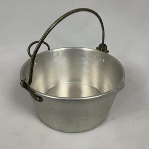 May include: A round, silver-coloured metal pot with a smooth interior and a tapered exterior. It features a dark, weathered metal swing handle attached to the sides with circular rivets and metal loops, allowing the handle to pivot freely.