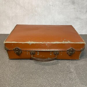 May include: A rectangular, burnt-orange suitcase featuring a worn, textured exterior with visible signs of age and surface wear. The case includes a central handle, two metallic latches on the front, and reinforced metal corners for added durability.