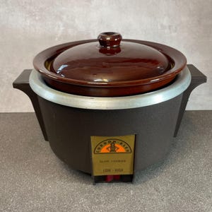 May include: A dark brown slow cooker with a matching dark brown ceramic lid and a silver-toned rim. The front features a rectangular label with the text CORDON BLEU SLOW COOKER LOW-HIGH and a graphic of a chef. The base has two side handles.