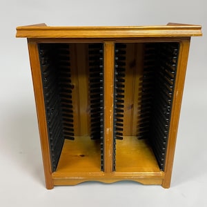 May include: A wooden CD storage cabinet in a light brown finish. It features two vertical compartments with black plastic dividers. The top and base are rectangular, and the sides are angled. The cabinet is designed to hold CDs.