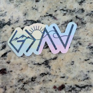 May include: A holographic sticker with a rainbow color scheme. The sticker features a mountain, sun, and wave design with the word "Grateful" written in black.