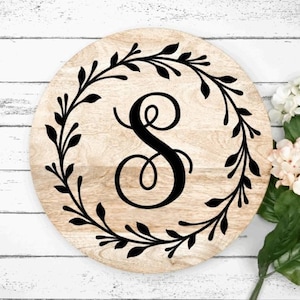 May include: A round wooden sign with a black wreath of leaves and the letter "S" in a decorative script font.