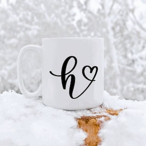 May include: White ceramic mug with a black cursive letter "h" and a small heart next to it. The mug is sitting on a snowy surface.