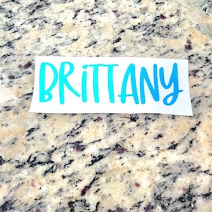 May include: A white vinyl sticker with the name "Brittany" written in a blue cursive font.