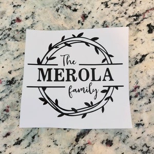 May include: A white sign with black text that reads "The Merola family" in a decorative font. The text is surrounded by a wreath of leaves.