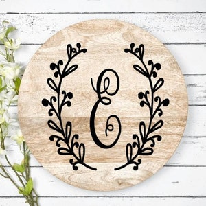 May include: A round wooden serving tray with a black floral wreath and the letter "E" in the center.